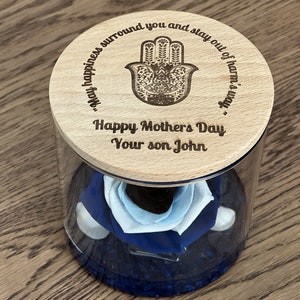 May include: A glass jar with a wooden lid containing a blue and white rose. The lid is engraved with a hamsa hand symbol and the text "May happiness surround you and stay out of harm's way."  The lid also has the text "Happy Mother's Day Your son John".