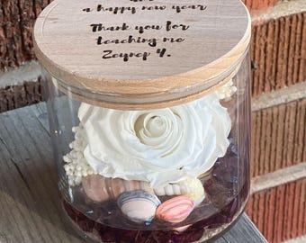 May include: A clear glass jar with a wooden lid engraved with the text: Happy holidays and a happy new year. Thank you for teaching me Zeynep 4. Inside the jar is a large white rose, small white filler flowers, seashells, and dark purple stones.