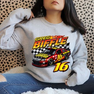 May include: A light grey sweatshirt with a graphic of a race car. The text reads "GREG BIFFLE" in yellow and red flames. The car is red and black with the number "16" and "NATIONAL GUARD" branding.