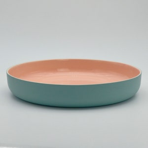 Może przedstawiać: Okrągła, płytka miska do serwowania z matowym wykończeniem. Zewnętrzna strona jest w stonowanym kolorze morskim, a wnętrze w delikatnym brzoskwiniowym. Misa przeznaczona jest do serwowania potraw i ma prostą, nowoczesną estetykę.