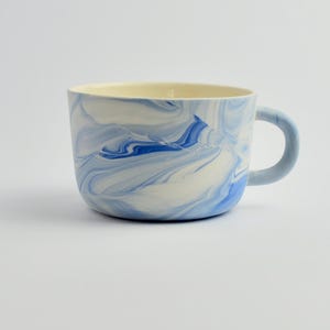 Puede incluir: Una taza de cerámica blanca con asa negra y el texto "But First, Coffee" en negrita negra. La taza se encuentra sobre un fondo borroso, sugiriendo un ambiente acogedor.