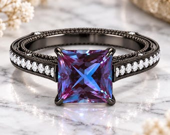 Può includere: Un anello in metallo nero con una pietra preziosa a taglio quadrato che cambia colore tra tonalità blu e viola. La fascia è impreziosita da piccole pietre rotonde trasparenti e presenta una texture a perline lungo i bordi.