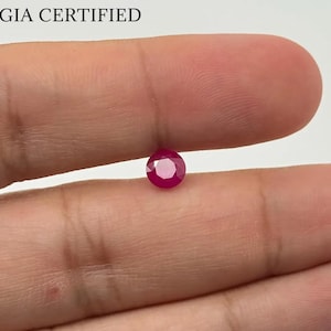 Puede incluir: Una gema de rubí rojo oscuro facetada. La gema es redonda con una base puntiaguda. El texto "GIA CERTIFIED" está impreso encima de la gema. El rubí se sostiene entre dos dedos.