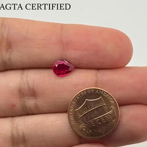 Può includere: Una gemma rossa vibrante a forma di pera accanto a una moneta da un centesimo. La gemma è di un colore rosso intenso e ricco. La moneta è color rame e mostra le parole "ONE CENT" e "UNITED STATES OF AMERICA". Il testo "AGTA CERTIFIED" è in alto.