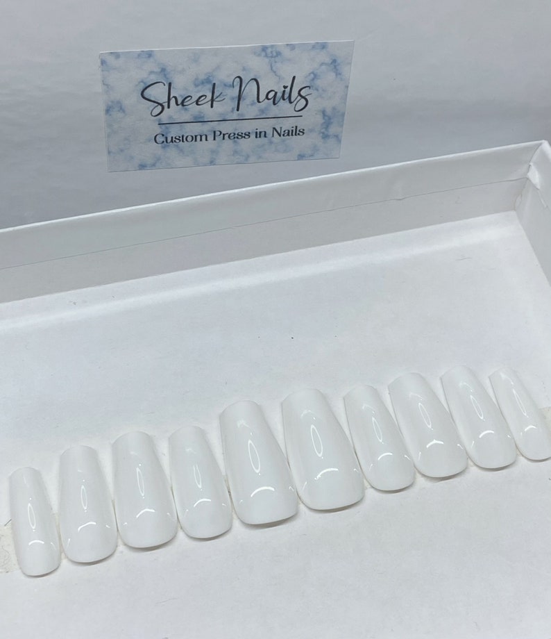 Pu&ograve; includere: Un set di dieci unghie finte bianche lucide disposte in fila all'interno di una scatola bianca. Sopra le unghie, un cartoncino rettangolare presenta un motivo a marmo blu con il testo Sheek Nails Custom Press in Nails stampato in inchiostro nero.