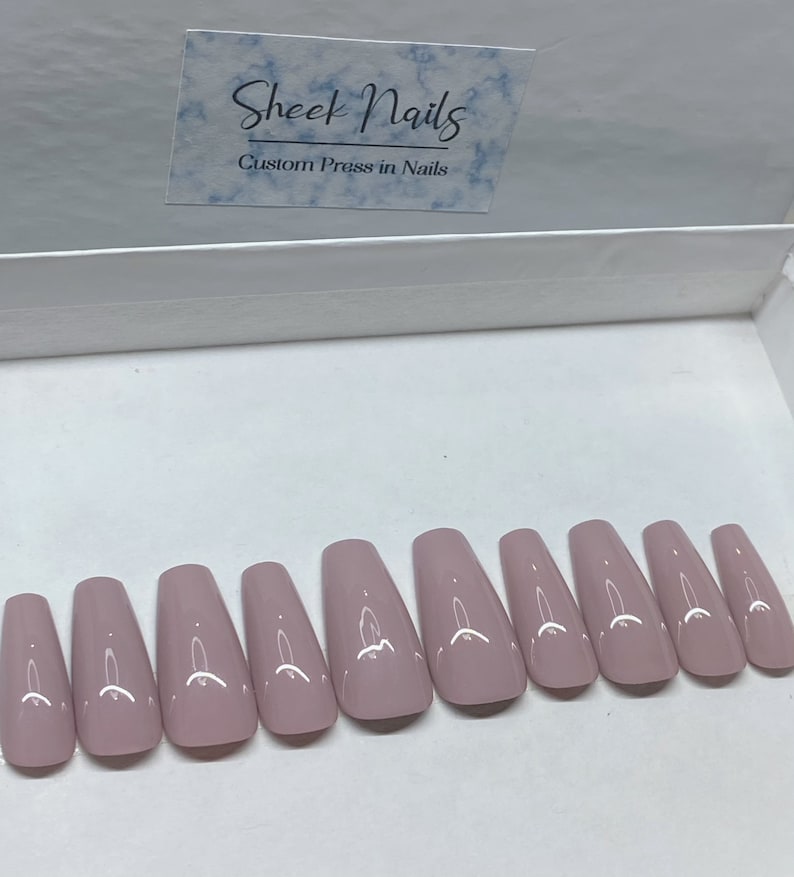 Pu&ograve; includere: Un set di dieci unghie finte lucide color malva disposte in fila all'interno di una scatola bianca. In alto &egrave; visibile un cartoncino rettangolare con motivo marmorizzato blu, con il testo Sheek Nails e Custom Press in Nails in nero.