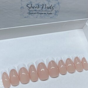May include: A set of custom press-on nails in a white box. The nails are oval-shaped with a natural pink base and a white French tip. The box has a label that reads "Sheek Nails Custom Press in Nails."