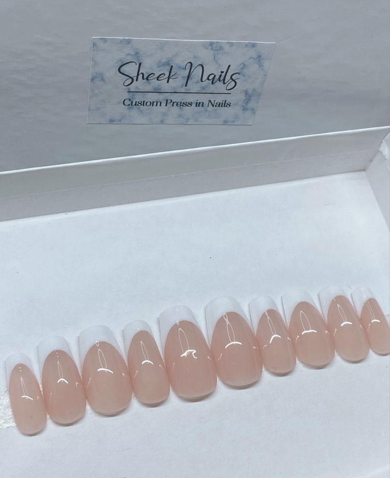 May include: A set of custom press-on nails in a white box. The nails are oval-shaped with a natural pink base and a white French tip. The box has a label that reads "Sheek Nails Custom Press in Nails."