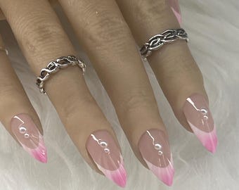 May include: Almond-shaped press-on nails featuring a French manicure style with a soft pink gradient tip, accented by small white pearls near the cuticle. Two silver-toned metal rings with a textured, chain-link design are worn on the fingers.