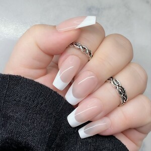May include: A hand showing French manicure false nails. The nails are rectangular with a sheer pink base and a white tip. Two silver rings are on the fingers.