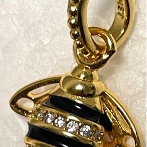 May include: A gold bee charm with black enamel stripes and clear crystal accents. The charm has a loop at the top for attaching to a bracelet or necklace. The charm is engraved with the word "Pandora".