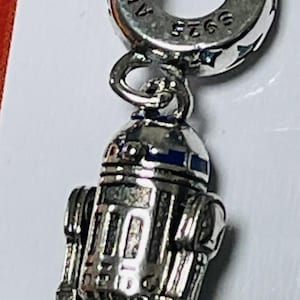 May include: A silver charm featuring a detailed depiction of R2-D2, a popular character from the Star Wars franchise. The charm is attached to a silver Pandora charm bracelet. The charm is marked with the text "Pandora 925 ALE."
