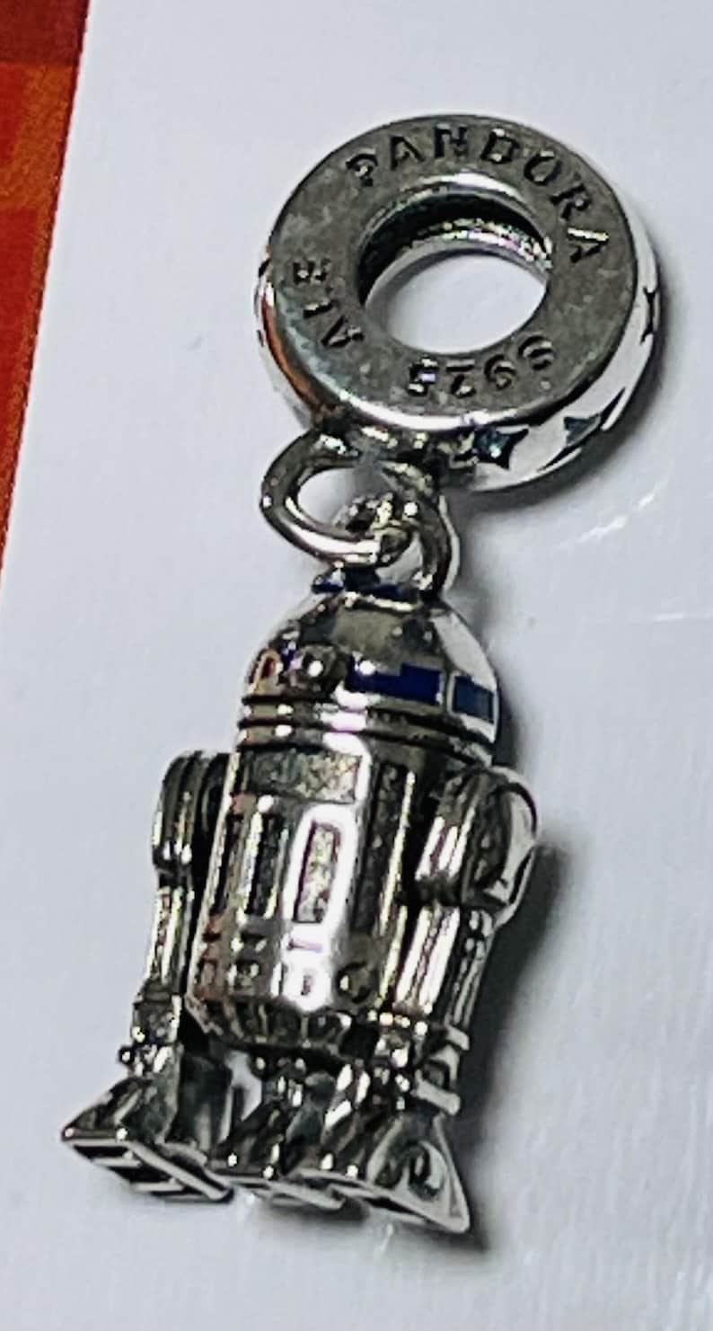 May include: A silver charm featuring a detailed depiction of R2-D2, a popular character from the Star Wars franchise. The charm is attached to a silver Pandora charm bracelet. The charm is marked with the text "Pandora 925 ALE."