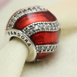Puede incluir: Un charm Pandora rojo y plateado con el nombre de la marca visible. El charm esférico presenta rayas de esmalte rojo y detalles plateados. Las secciones plateadas están engastadas con pequeñas piedras transparentes.