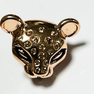 Puede incluir: Un amuleto de animal de tono dorado con un patrón de manchas y ojos negros. El amuleto tiene la forma de la cabeza de un leopardo.