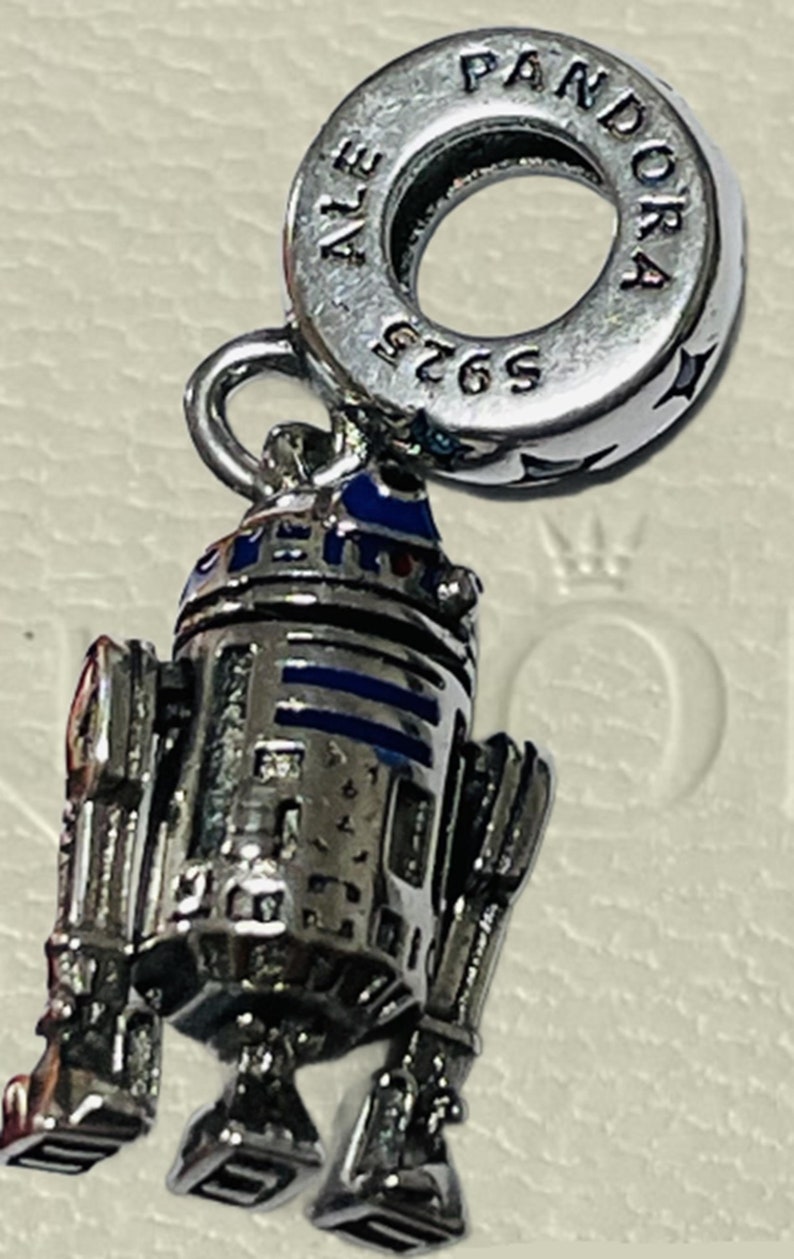 May include: A silver charm featuring a detailed R2-D2 robot with blue and white accents. The charm is attached to a silver Pandora charm bracelet with the text "Pandora" and "ALE 5265" engraved on the back.