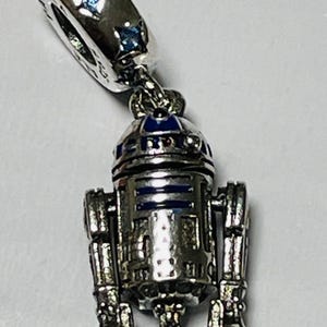 May include: Silver charm featuring a detailed R2-D2 robot with blue and silver accents. The charm has a small loop at the top for attaching to a bracelet or necklace.