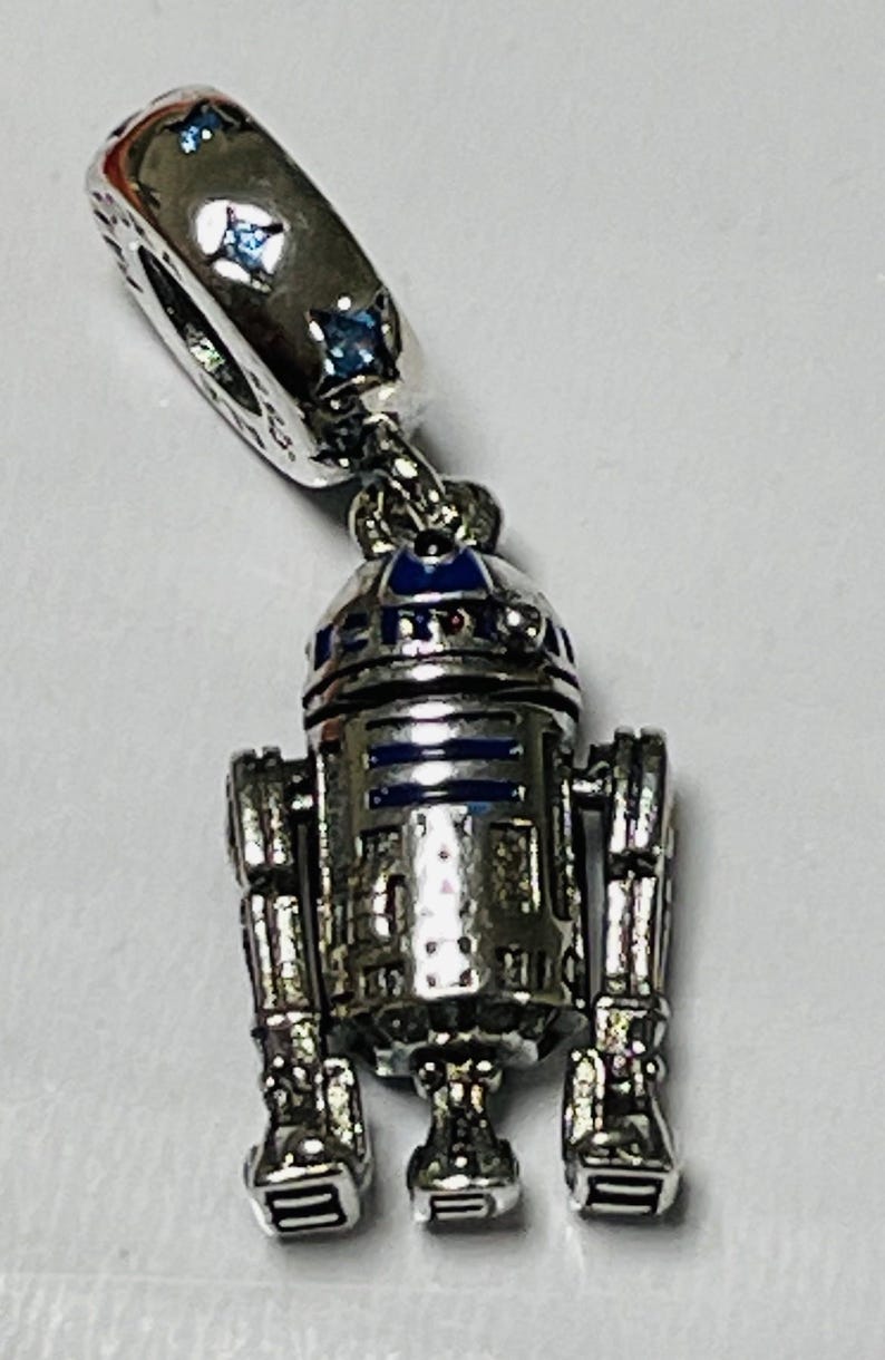 May include: Silver charm featuring a detailed R2-D2 robot with blue and silver accents. The charm has a small loop at the top for attaching to a bracelet or necklace.