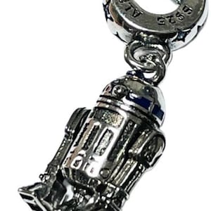 May include: A silver charm featuring a detailed depiction of R2-D2, a popular character from the Star Wars franchise. The charm is attached to a silver Pandora charm bracelet with the Pandora logo and 925 ALE inscription.