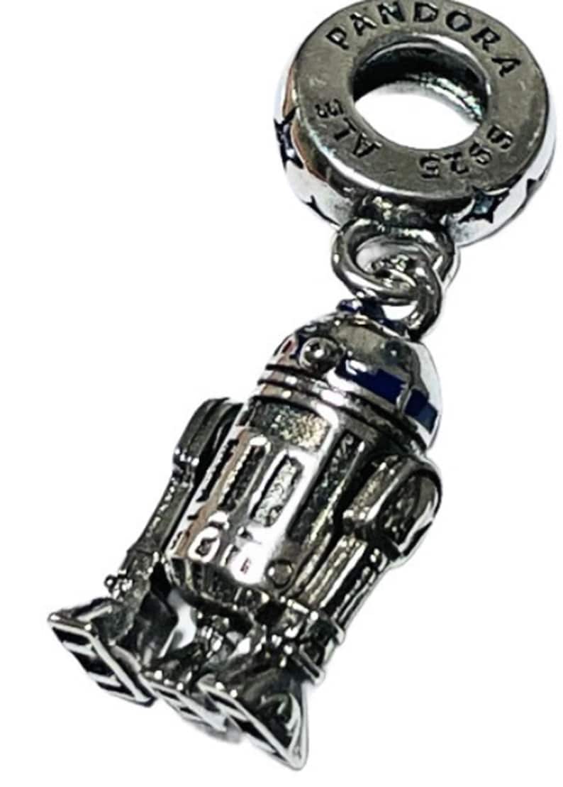 May include: A silver charm featuring a detailed depiction of R2-D2, a popular character from the Star Wars franchise. The charm is attached to a silver Pandora charm bracelet with the Pandora logo and 925 ALE inscription.
