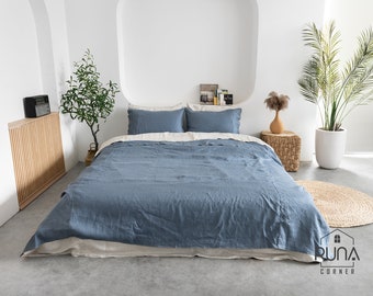 May include: A blue linen duvet cover with two white pillowcases on a bed in a modern bedroom. The bed is made with a white linen sheet and has a wooden nightstand with a radio on it. There is a potted plant and a vase with dried flowers on the nightstand.