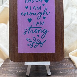 May include: A wooden sign with a purple background and teal lettering. The sign reads "I am loved, I am enough, I am strong" with hearts and a leafy branch.