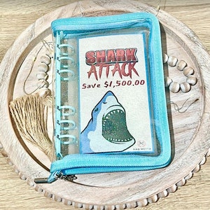 May include: A blue zippered binder with a clear plastic front. The front features a shark with glitter and the text "SHARK ATTACK Save $1,500.00".