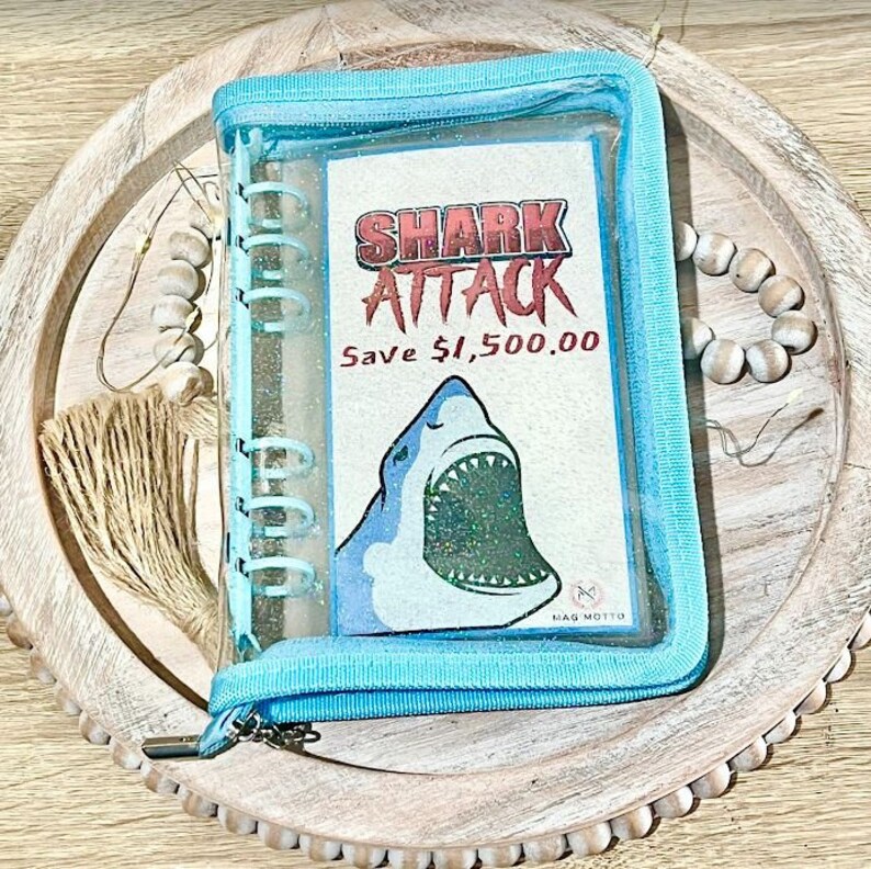 May include: A blue zippered binder with a clear plastic front. The front features a shark with glitter and the text "SHARK ATTACK Save $1,500.00".