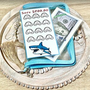 May include: A blue zippered pouch with a clear plastic window containing a shark tooth chart and a $1000 bill. The chart has a shark graphic and the text "Save $200.00".