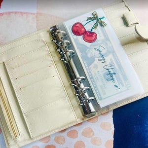 May include: A cream-colored leather planner with a clear plastic pocket containing a dollar bill and a cherry graphic. The pocket is labeled "Savings Challenge #1".