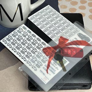 May include: A black leather planner with a clear plastic pocket containing a sticker sheet with car icons and a sticker sheet with car keys. The planner has a red ribbon and the text "New Car Savings Challenge" is visible.