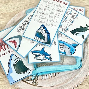 May include: A set of colorful shark-themed educational flash cards with different amounts of money on each card. The cards are in a clear plastic pouch with a blue zipper.