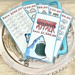 May include: A set of five colorful cards with shark-themed illustrations and text that reads "Save $100.00", "Save $200.00", "Save $300.00", "Save $400.00", and "Save $500.00". The cards are arranged in a stack with the largest card on top. The cards are in a clear plastic pouch.
