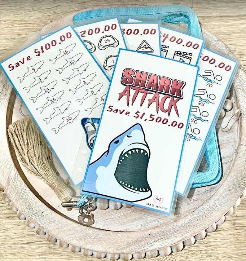 May include: A set of five colorful cards with shark-themed illustrations and text that reads "Save $100.00", "Save $200.00", "Save $300.00", "Save $400.00", and "Save $500.00". The cards are arranged in a stack with the largest card on top. The cards are in a clear plastic pouch.