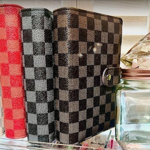 May include: Four different patterned hardcover journals with a checkered design. The journals are stacked on a clear acrylic shelf.