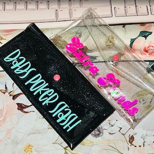 May include: Two rectangular pouches. One is black with glitter and the words "Dads Poker Stash" in teal. The other is clear with pink text that says "Sweethearts". Both have a pink snap closure.