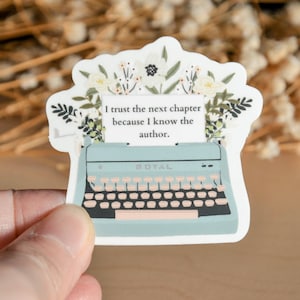 May include: A blue typewriter with a white paper insert that reads "I trust the next chapter because I know the author."  The typewriter is surrounded by white flowers and green leaves.