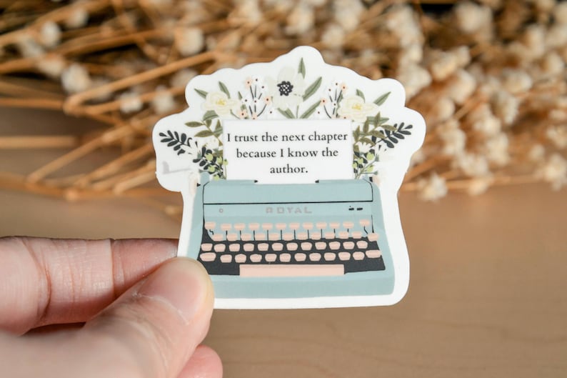May include: A blue typewriter with a white paper insert that reads "I trust the next chapter because I know the author."  The typewriter is surrounded by white flowers and green leaves.