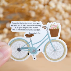 Peut inclure: Un autocollant découpé représentant un vélo bleu clair avec une selle marron et un panier marron rempli de fleurs roses. Le texte indique : Trust in the Lord with all your heart and lean not in your own understanding; in all your ways submit to him, and he will make your paths straight. -Proverbs 3:5-6.