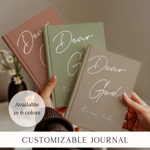 May include: Three hardcover journals in muted terracotta, sage green, and beige. Each journal features the white script text 'Dear God' and 'It's me, Kate' on the front cover. The journals are held in hands, showcasing their solid color design.