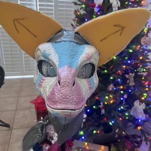 May include: A partially finished, grey, pink, and blue furry mask with yellow foam ears. The mask has a textured surface and is on a stand. The mask is in front of a Christmas tree with colorful lights.