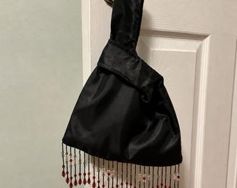 May include: A black satin wristlet handbag featuring a single integrated handle and a bottom fringe composed of clear and deep red teardrop-shaped beads. The fabric has a smooth, slightly reflective finish, and the beaded fringe hangs in a scalloped pattern.