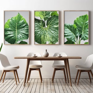 May include: Three framed botanical prints featuring close-up images of large, vibrant green leaves with detailed veining. The prints are displayed above a wooden dining table with white chairs, creating a modern, nature-inspired interior design.