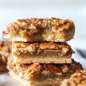 Op de afbeelding: Een stapel van drie pecannoten taartrepen met een gouden korst en een topping van gekarameliseerde pecannoten. De repen zijn gerangschikt op een wit oppervlak.