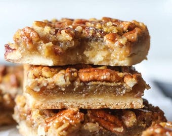 May include: A stack of three pecan pie bars with a golden crust and a topping of caramelized pecans. The bars are arranged on a white surface, with a soft focus background.