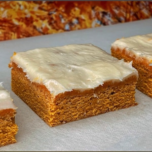 May include: Three square-shaped baked goods with a light-colored frosting. The cake has a golden-brown color and a textured appearance. The frosting is thick and creamy, covering the top of each piece. The baked goods are arranged on a white surface.