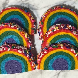 Op de afbeelding: Zes regenboogkleurige koekjes, in een halve cirkel gesneden, tonen een levendig kleurenspectrum. De koekjes zijn bedekt met kleurrijke hagelslag, wat textuur toevoegt. Een leuke en feestelijke traktatie.