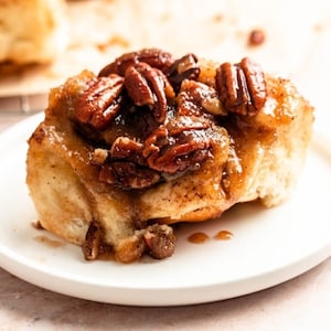 Könnte beinhalten: Nahaufnahme eines Pecan-Sticky-Buns mit einem süßen, klebrigen Guss und Pecan-Stücken darauf. Der Bun liegt auf einem weißen Teller.