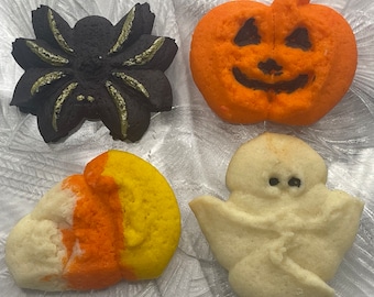 May include: Four Halloween-themed cookies: a black spider with gold accents, an orange jack-o'-lantern, a candy corn shape, and a white ghost. The cookies are arranged on a textured, white surface.