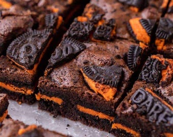 Könnte beinhalten: Nahaufnahme von Schokoladenbrownies mit orangefarbener Füllung und dunklen Schokoladenkeksstücken. Die Brownies sind in Quadrate geschnitten und auf einer weißen Oberfläche angeordnet. Das Bild hebt die Textur und den Farbkontrast des Desserts hervor.